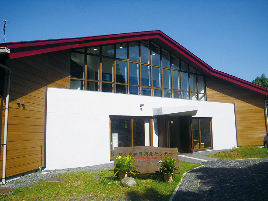 A front view of the Yakushima World Heritage Conservation Center. The building has two floors with a gable roof, and a wide expanse of dormer windows at the front above the entrance. The wooden sign in front of the building reads 'Yakushima World Heritage Conservation Center'.