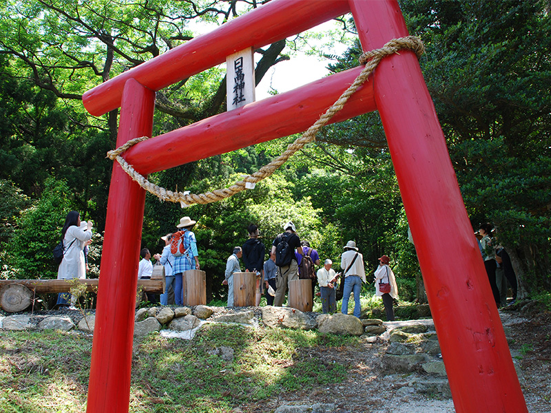 A village tour in progress. Tourists receive explanations from a guide within the grounds of a shrine in the village. The red torii gate of the shrine can be seen in the foreground. Tourists chatting and taking photos.