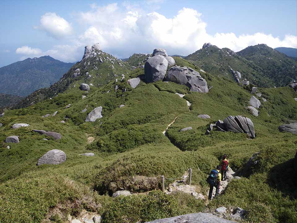 A hiking trail providing an experience of the pristine natural environment of the mountains of Yakushima Island. Two climbers walk along a narrow trail on a mountain ridge. The mountains are dotted with many large, craggy boulders.