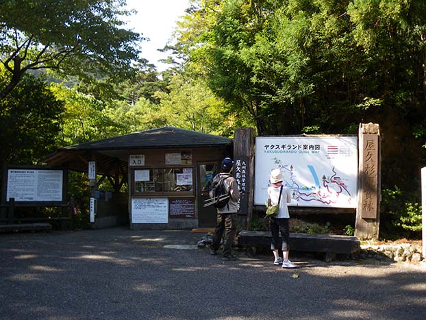 The Yakusugi Land Entrance. A large sign with a map of Yakusugi Land is shown in the right of the photo, and on the left is a small wooden administration building that serves as the entrance. Two tourists in front of the map.