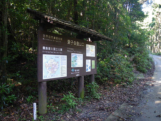 愛子岳歩道入口に設置された、木製の案内標識の写真。板面には、世界自然遺産地域、屋久島国立公園、愛子岳登山口といった文字が書かれており、数枚の地図や説明書きも貼られています。