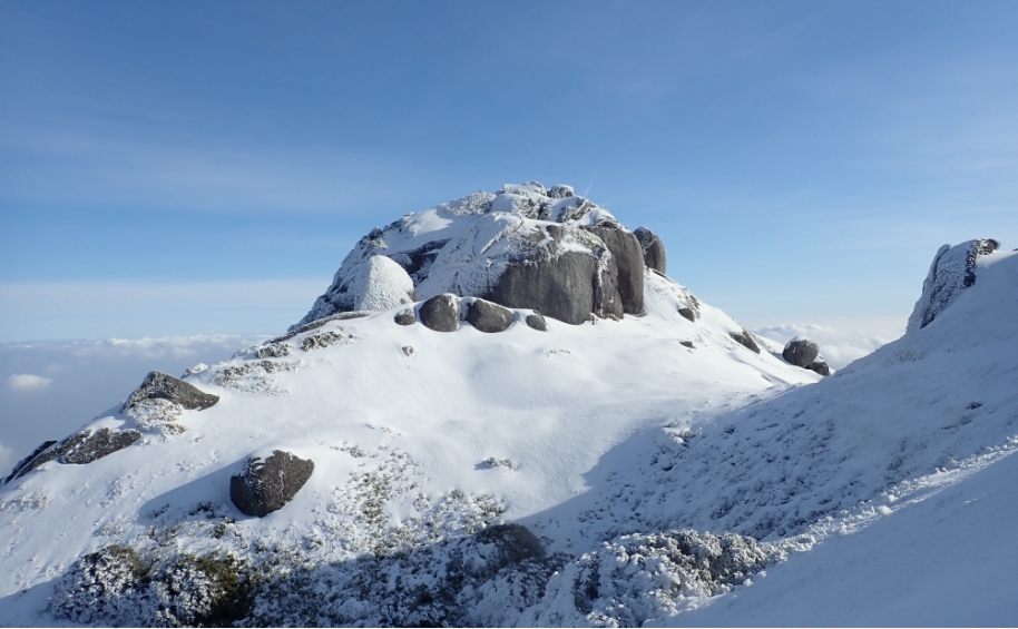 冬季山区照片。山峰高耸入云，纯白的雪覆盖着山体。然而，山顶的岩石上被风吹裹上一层薄雪。