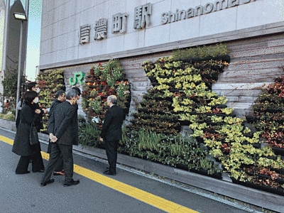 JR信濃町駅の壁に、様々な花や植物が色とりどりに飾られて、様々な絵を織りなしています。それらを壁の前で複数の人が眺めています。