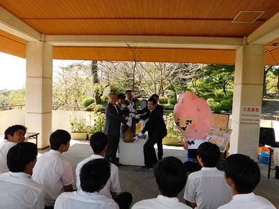 公園の休憩所で、学生達が話を聞いています。話をしているスーツを着た大人達は1本の木を持っています。横にはマスコットキャラクターもいます。