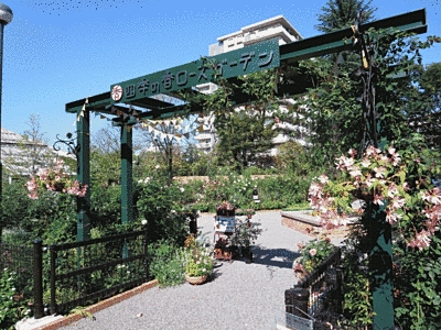 四季の香ローズガーデンと書かれた緑色の入り口の門があります。門の両サイドにはピンクの花や緑がいっぱいです。門をくぐった先には、花や木の他にベンチも見えます。