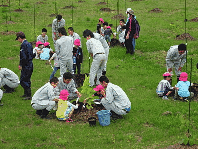 広い原っぱで植樹作業が行われています。大人の他に、ピンクの帽子をかぶった子供たちの姿もあります。