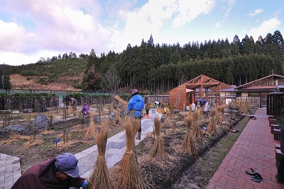 奥には山、林が見えます。手前の施設内の庭のような場所では、何人かが作業しており、藁のようなものを立てています。