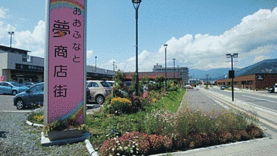 おおふなと夢商店街の大きなピンク色の看板が左側にあります。駐車場と道の間には木や花が植えられており、大変鮮やかです。