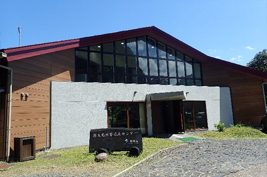 A front view of the Yakushima World Heritage Conservation Center. The building is characterized by a dark red triangular roof and the wide expanse of upper windows. The sign in front of the building reads 'Yakushima World Heritage Conservation Center'.