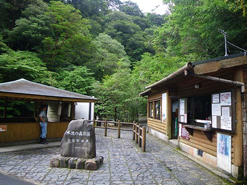 The Shirataniunsui-kyo Ravine Entrance. The wooden administration building on the right, a small rest area on the left, and a stone in the center noting the '100 Best Forest Water Sources'.