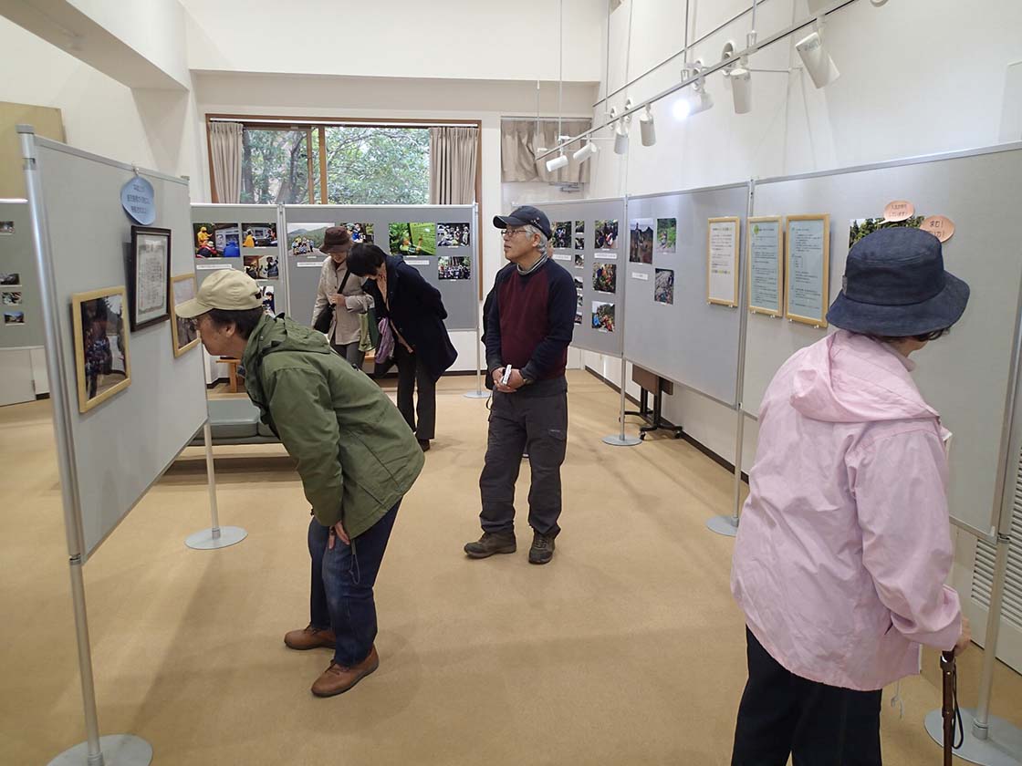 屋久島国立公園で活躍する屋久島国立公園パークボランティアの活動を紹介する、活動20周年記念活動写真展の様子。写真には、5名の来館者が見えます。身をかがめ、熱心に写真を見ている人もいます。