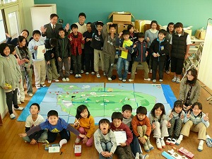 島内の小学校での実施風景で、屋久島・口永良部島の地図を中央に、小学生がぐるっと取り囲んだ集合写真。