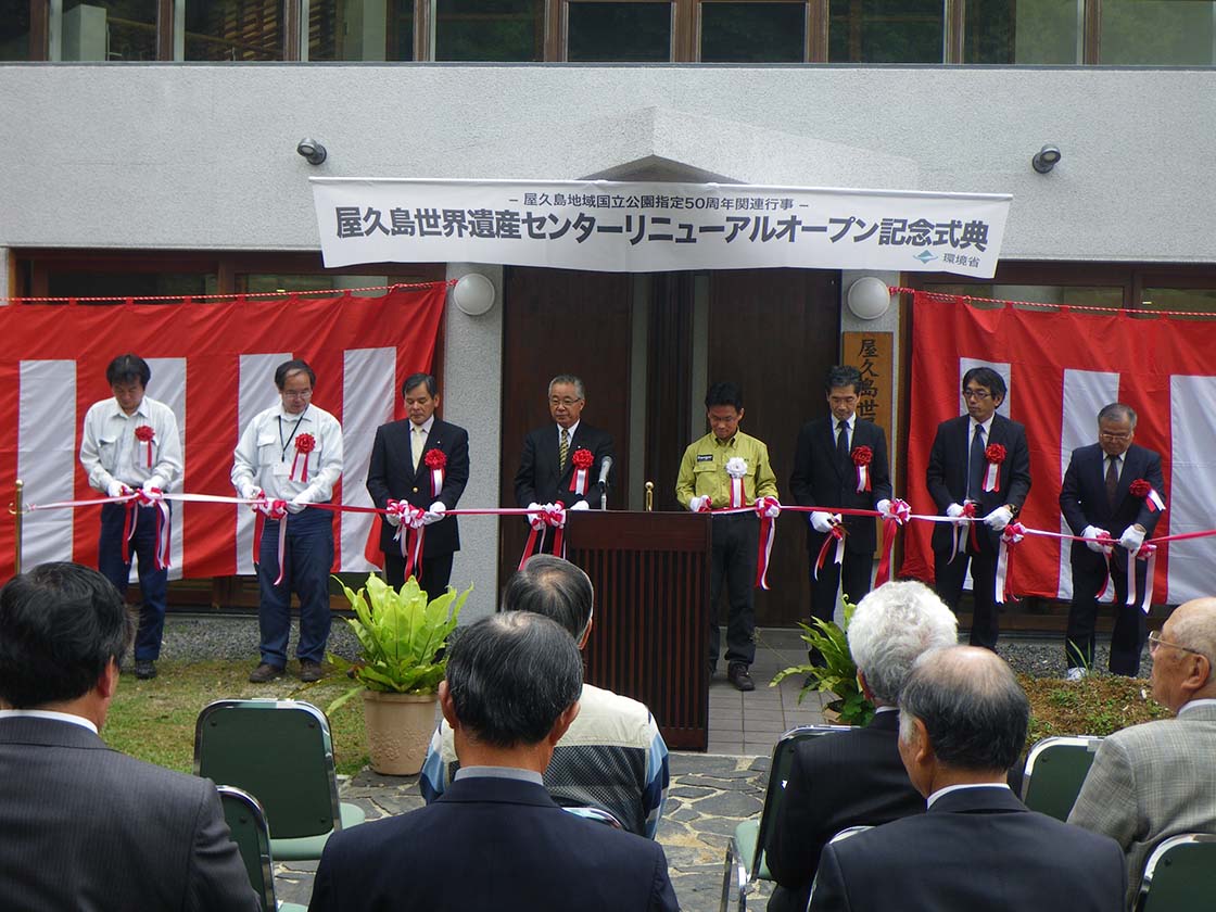 平成26年度に行った、屋久島世界遺産センターリニューアルオープン記念式典の様子。入口付近が紅白幕に覆われた建物を背に、胸に赤い花をつけた来賓者7名が、テープカットを行う瞬間です。