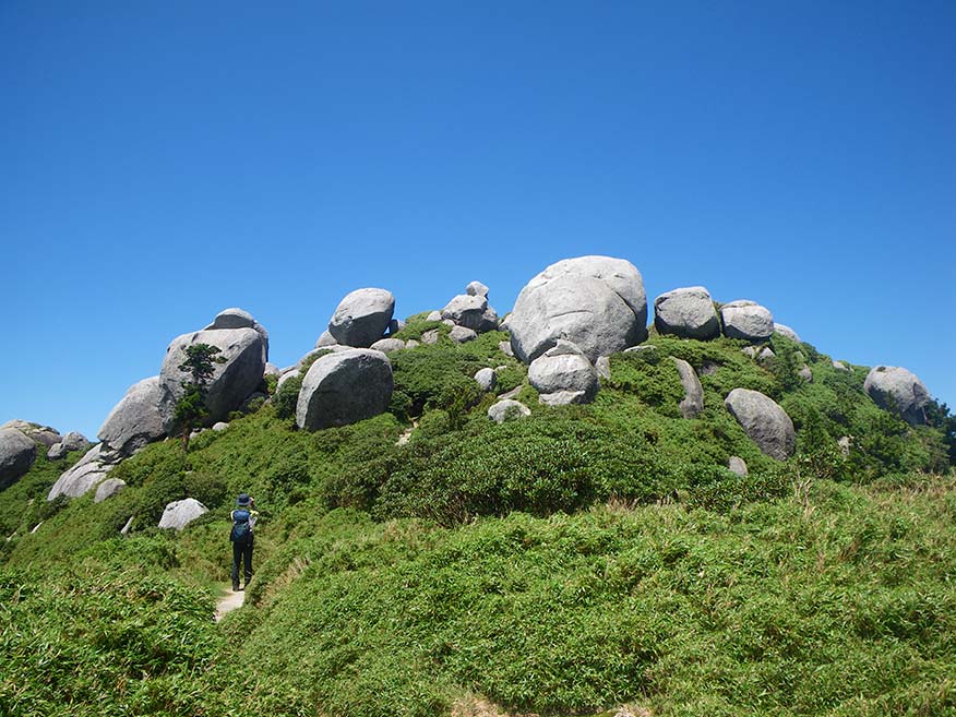 巨大な岩が、山の上に露出している平石岩屋の写真。一人の登山者が登山をしており、いかに巨大な石か、大きさがうかがいしれます。