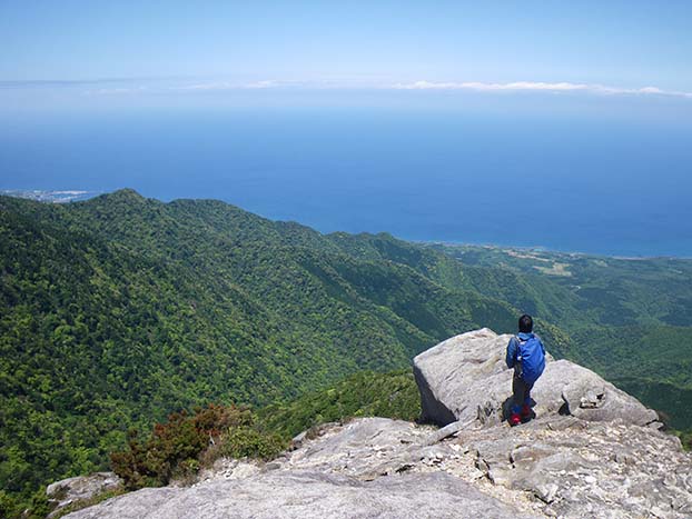 愛子岳山頂の写真。花崗岩の岩場になっており、一人の登山者が目の前に広がる屋久島北西部の山や海を眺めています。
